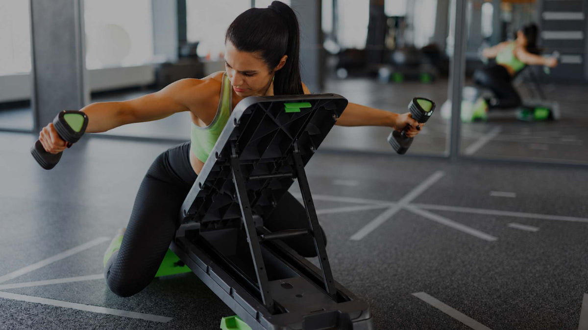 A woman in a gym kneels on the floor while using a black step platform with dumbbells, wearing a green sports tank top and black leggings.