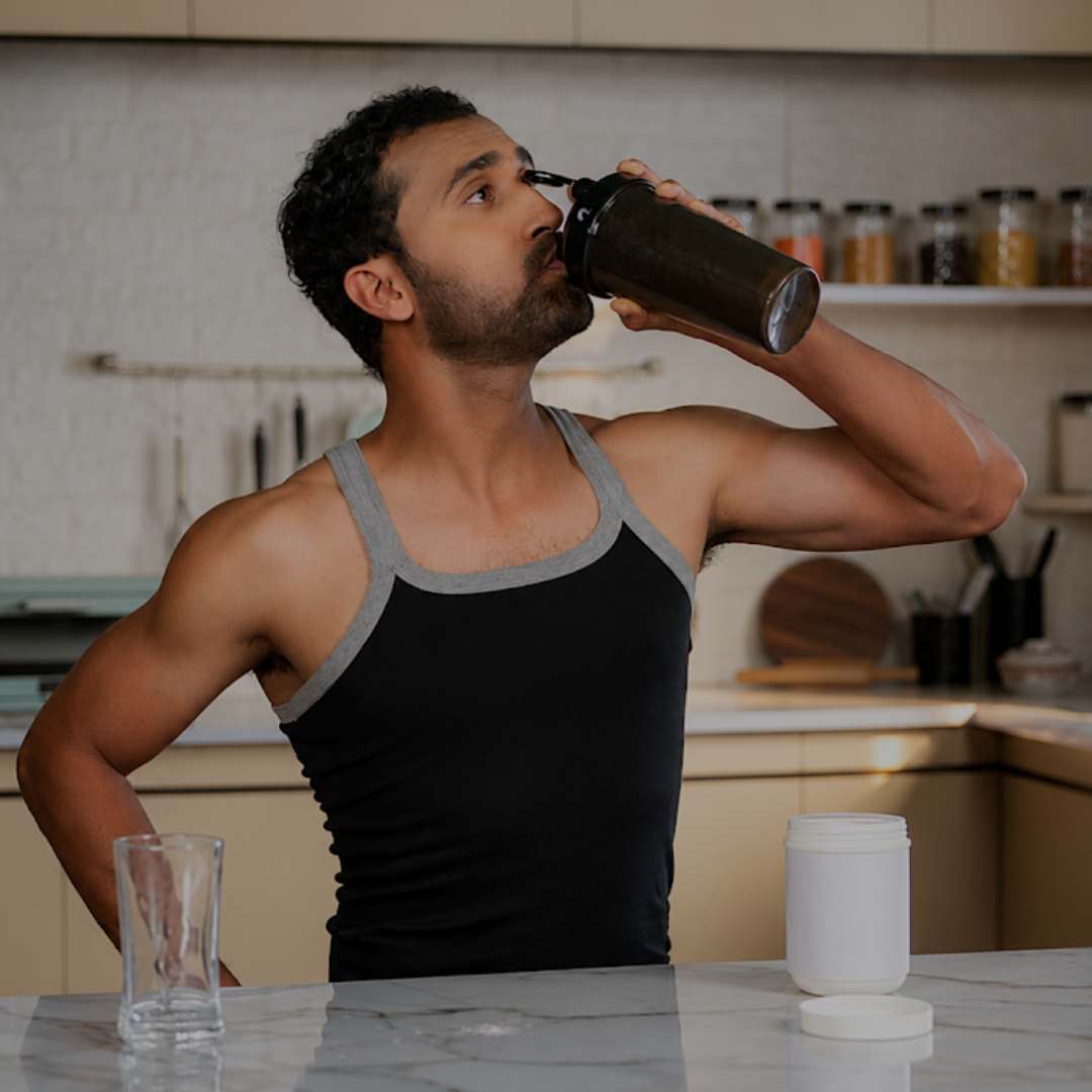 A man in a black and gray tank top drinks from a black shaker bottle on a kitchen counter, with a white supplement jar and a glass nearby.