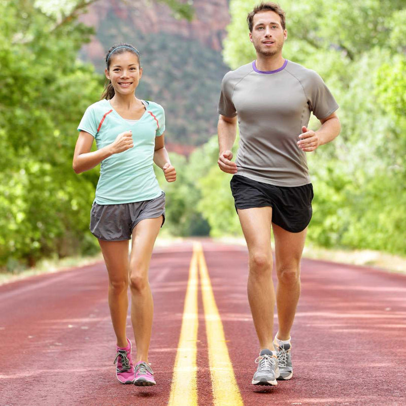 A man and a woman jogging side-by-side on a paved road with double yellow lines, surrounded by lush green trees and a rocky cliff in the background. The woman wears a light blue-green t-shirt and gray shorts, while the man wears a gray t-shirt and black shorts.