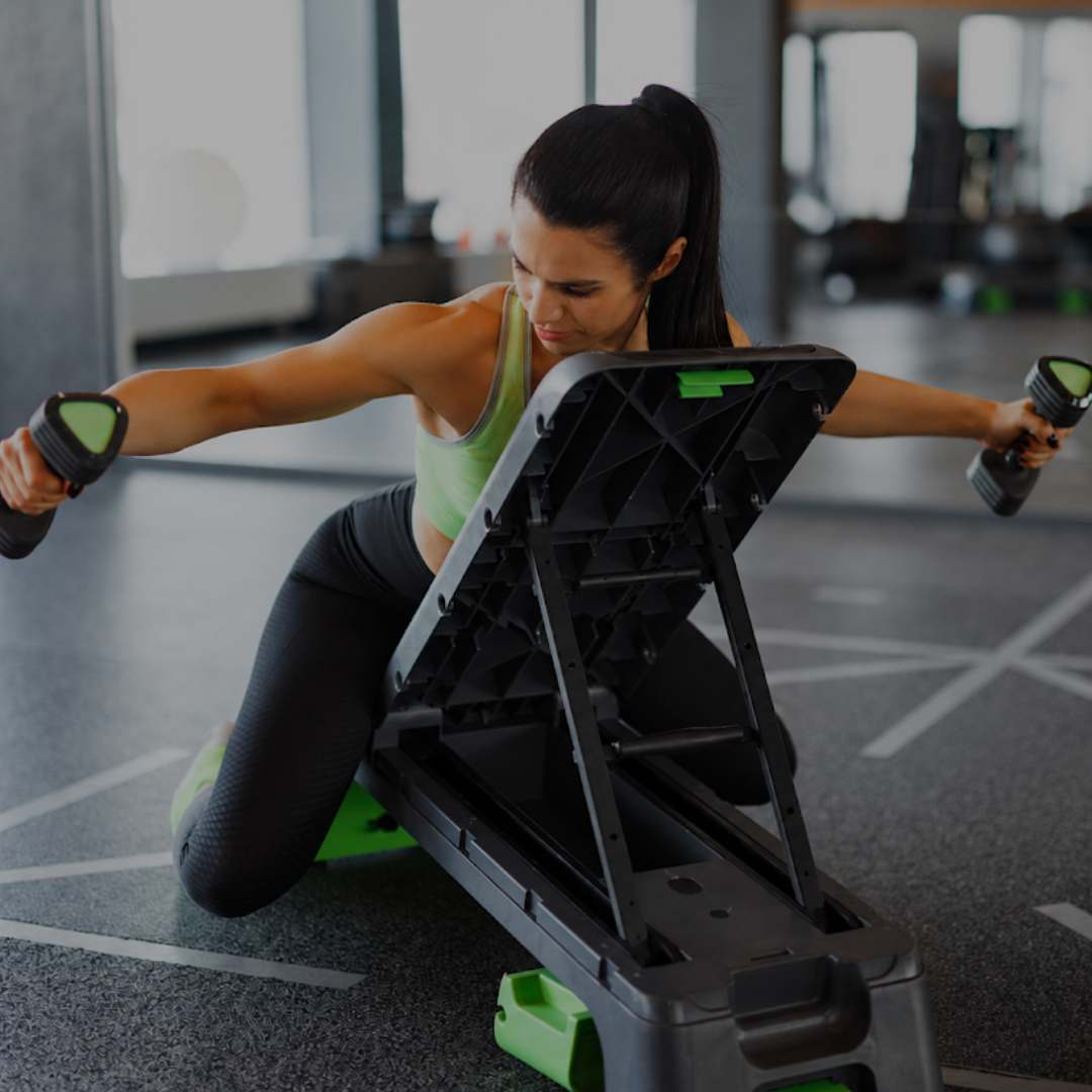 A woman in a gym kneels beside a large black and green fitness storage cart, holding dumbbells with green accents.