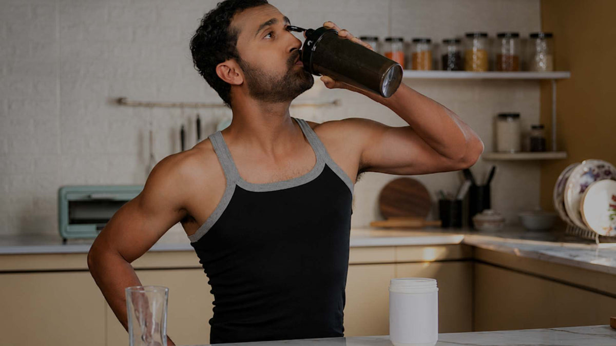 A man in a black and gray tank top drinking from a shaker bottle in a kitchen setting, with a white tub on the counter in front of him.