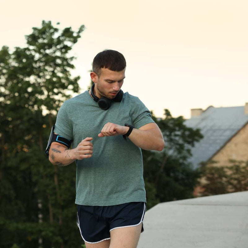 A man outdoors wearing a green athletic t-shirt and navy running shorts, looking down at a smartwatch on his wrist. He has a black armband and headphones around his neck, with trees and a building in the background.