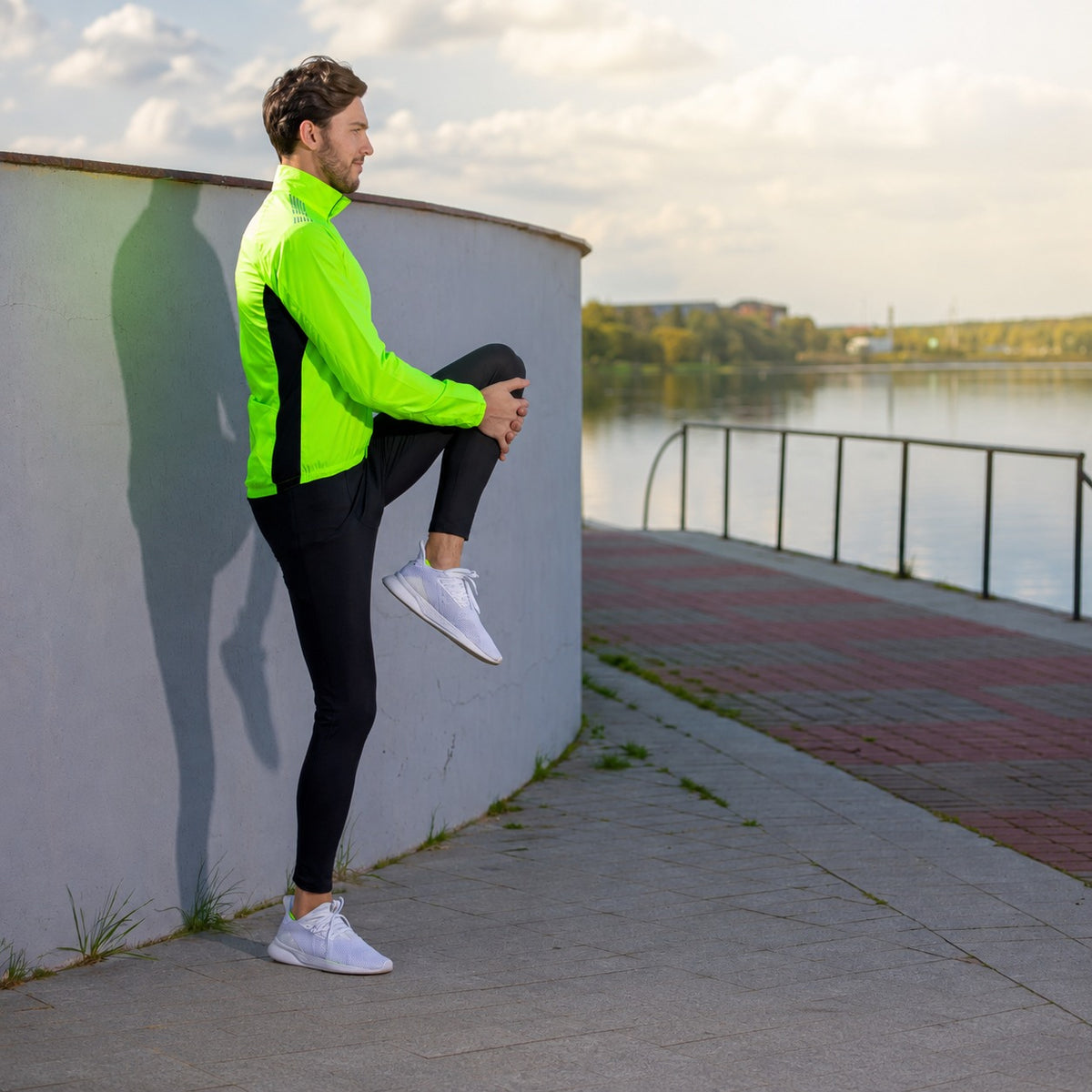 A man in a bright neon green and black athletic jacket, black leggings, and white sneakers stretches his leg against a curved gray wall next to a waterfront walkway.