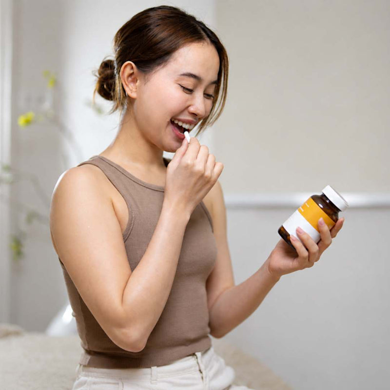 A woman in a brown tank top holds a brown supplement bottle and is shown taking a pill from her hand, set against a soft, neutral indoor background.